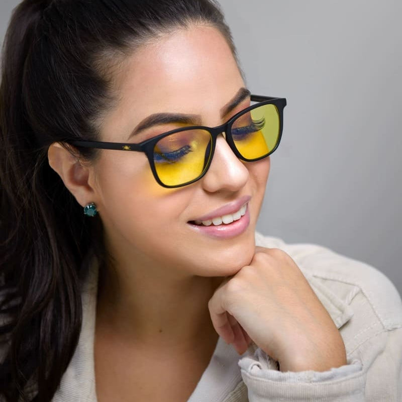 Una mujer con cabello oscuro recogido en una cola de caballo, sonriendo ligeramente mientras usa gafas anti luz azul con tintes amarillos y montura negra, y una camisa beige.