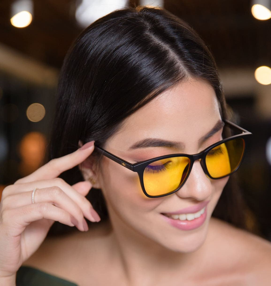 Una mujer con cabello negro liso, tocando el marco de sus gafas anti luz azul Bioptimizacion de montura negra y lentes tintados de amarillo, sonriendo suavemente con un top verde oscuro con hombros descubiertos y volantes en un ambiente suavemente iluminado con luces desenfocadas en el fondo.