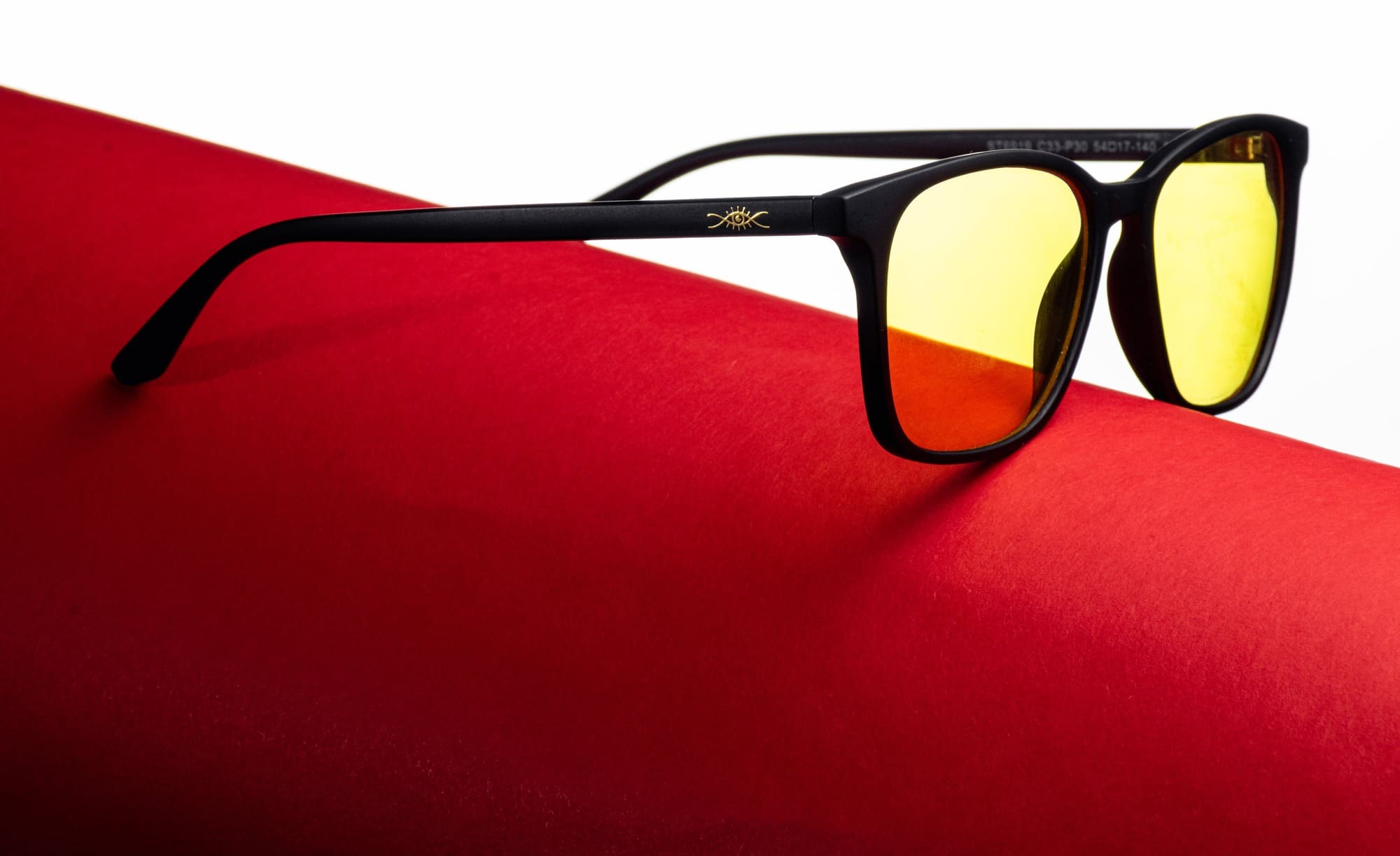 Un primer plano de unas gafas blue block de montura negra marca "bioptimizacion" y lentes tintados de amarillo, situadas sobre una superficie roja vibrante, con un pequeño logotipo similar al sol en la montura, mostrando un fuerte contraste de colores y un diseño elegante.