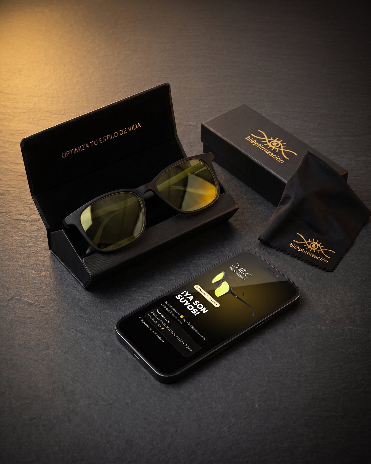 Lentes Alpha Clarity+ sobre su estuche rígido negro, junto al paño con logo dorado y la caja