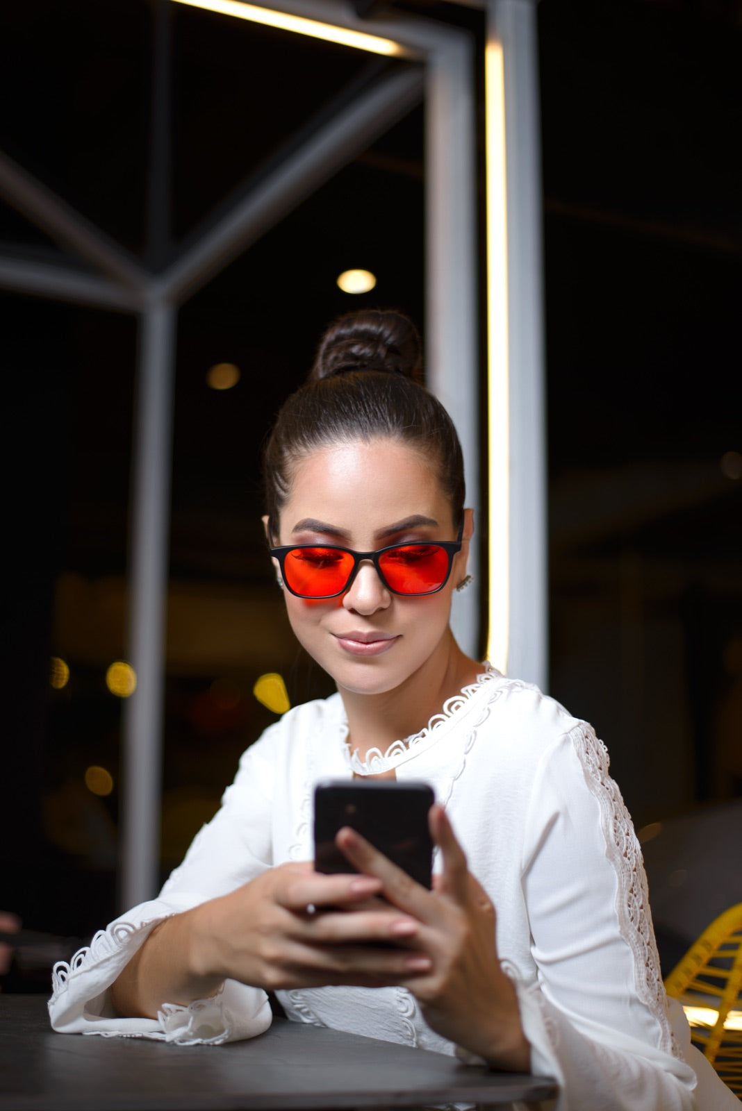 Mujer usando su celular de noche junto a una ventana con los lentes Alpha + Sueño puestos